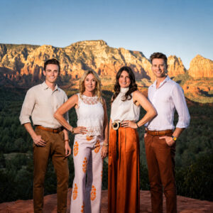 Four real estate agents from the Sedona Luxury Homes team smiling and posing for a professional group portrait. They stand confidently outdoors against a scenic backdrop of Sedona's iconic red rock formations and green desert vegetation under bright, golden-hour sunlight.