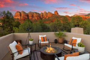 Rooftop patio with outdoor rocking chairs and a flaming fire pit overlooking lush green treetops and red rock mountains at sunset
