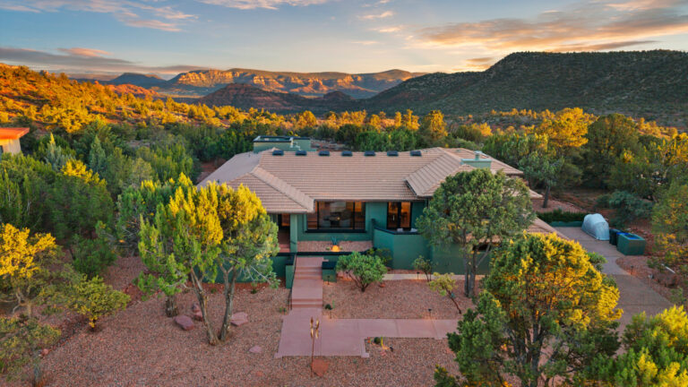 High aerial view of a single-story desert home nestled among evergreen trees with red rock mountain vistas glowing at sunset